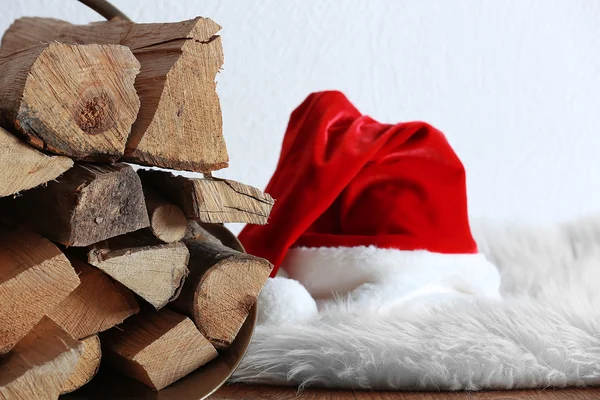Basket with firewood and Santa Claus hat on floor against white wall, close up — Stock Photo, Image