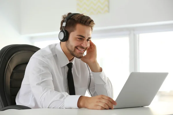 Schöner junger Mann, der mit Kopfhörern Musik hört und im Büro am Laptop arbeitet — Stockfoto