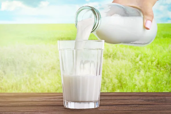 Female hand pouring milk into glass against blurred nature background. Dairy concept. — ストック写真