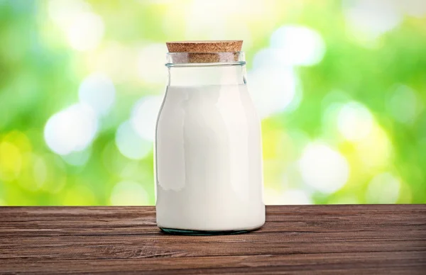 Glass bottle of milk on wooden table against blurred nature background. Dairy concept. — Stock fotografie