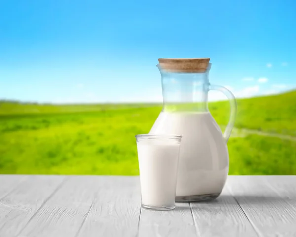 Vidro e jarro com leite na mesa de madeira branca contra fundo natureza turva. Conceito de leiteria . — Fotografia de Stock