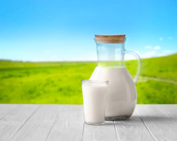 Verre et cruche avec du lait sur table en bois blanc sur fond de nature floue. Concept laitier . — Photo