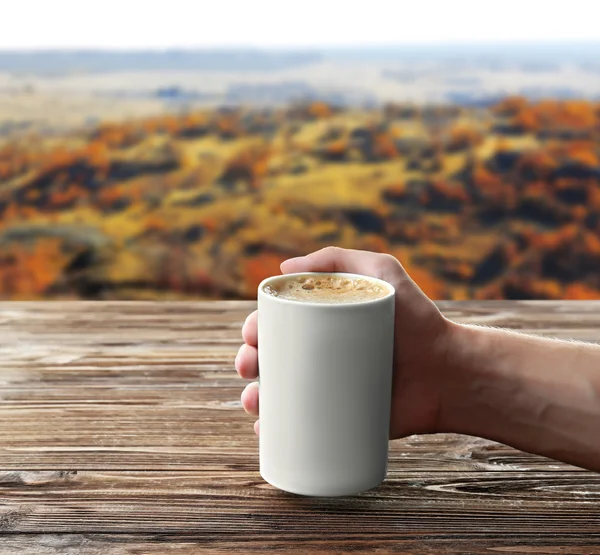 Mão de homem com xícara de café na mesa de madeira contra fundo paisagem — Fotografia de Stock