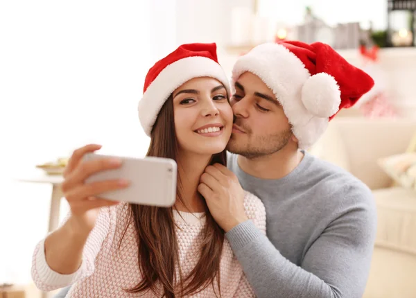 Pareja joven en Santa Claus sombreros haciendo selfie en el interior —  Fotos de Stock