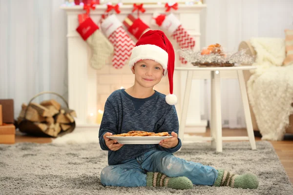 Aranyos kisfiú Santa kalap tányér finom sütit otthon — Stock Fotó