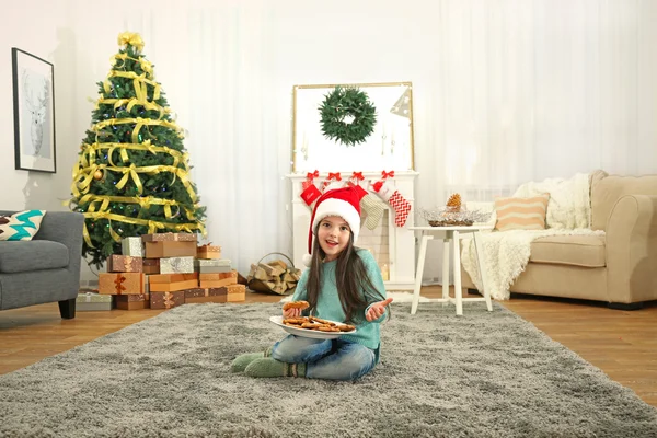 Το χαριτωμένο μικρό κορίτσι το καπέλο Santa με πλάκα από νόστιμα μπισκότα στο σπίτι — Φωτογραφία Αρχείου