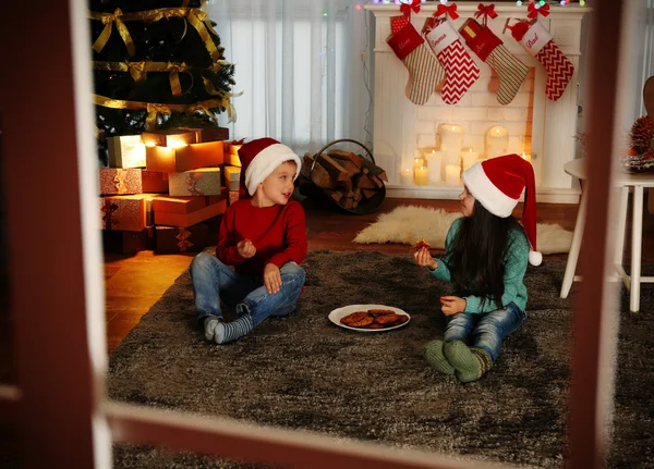 Bonito crianças em chapéus de Santa comer deliciosos biscoitos em casa, vista através da janela — Fotografia de Stock