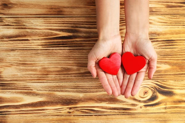 Mãos femininas segurando corações vermelhos no fundo de madeira — Fotografia de Stock