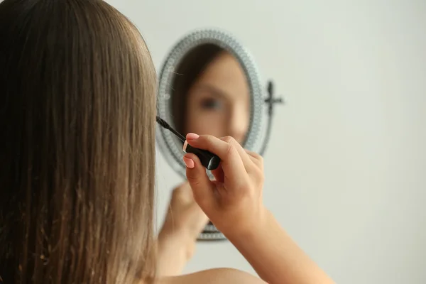Όμορφη νεαρή γυναίκα κοιτάζοντας στον καθρέφτη, κατά την εφαρμογή του μακιγιάζ — Φωτογραφία Αρχείου