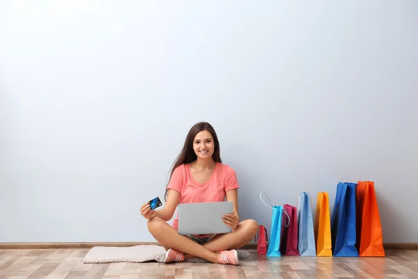 Wanita muda yang bahagia dengan tas belanja berwarna-warni dan laptop duduk di lantai — Stok Foto