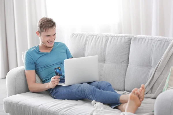 Молодой человек с ноутбуком сидит на диване и делает онлайн-покупки — стоковое фото