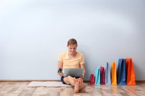 Happy young man with colorful shopping bags and laptop sitting on floor — Stock Photo, Image