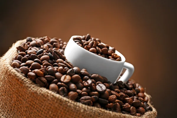Sack with roasted coffee beans and white cup on dark background, close up view — Stock Photo, Image