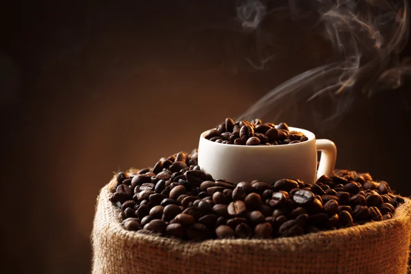 Sack with roasted coffee beans and white cup on dark background, close up view — Stock Photo, Image