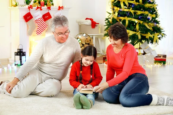 Bambina e i suoi nonni leggono un libro in salotto decorato per Natale — Foto Stock