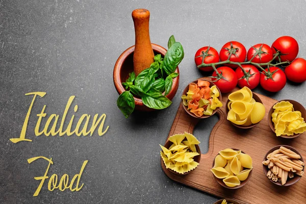 Bowls with different pasta on wooden cutting board. Text ITALIAN FOOD on gray background. Traditional meal concept. — Stock Photo, Image