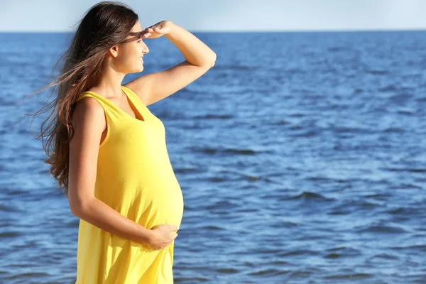 Νεαρή έγκυος γυναίκα στο κίτρινο φόρεμα στέκεται στην παραλία — Φωτογραφία Αρχείου
