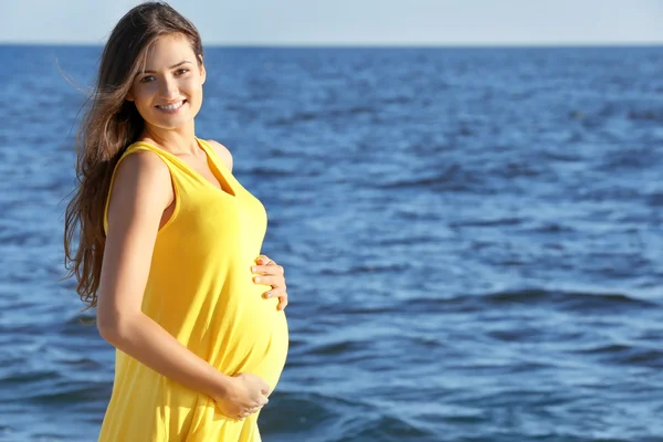 Wanita hamil muda dengan gaun kuning berdiri di pantai — Stok Foto