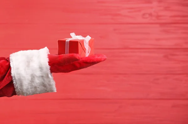 Рождественская идея. Рука Санта-Клауса держит подарочную коробку на красном деревянном фоне — стоковое фото