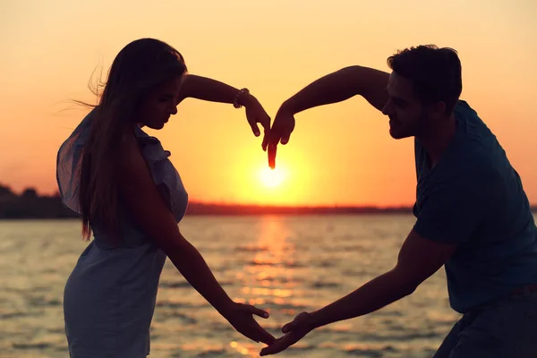 Pareja joven enamorada cogida de la mano en forma de un gran corazón en la playa al atardecer — Foto de Stock