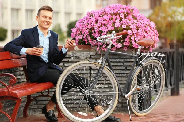 Coffee-to-go concept. Young man with bicycle and cup of coffee sitting on the bench
