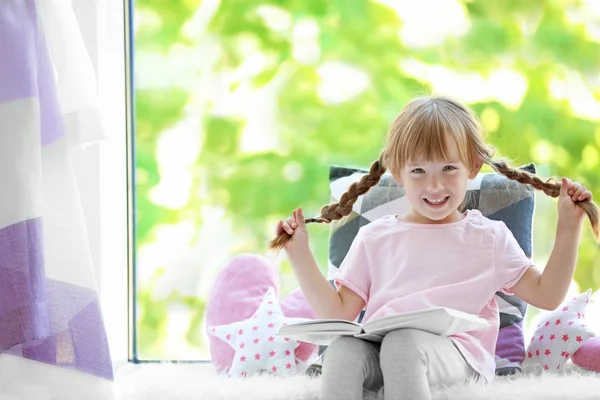 Vacker liten flicka med boken på fönsterbrädan — Stockfoto