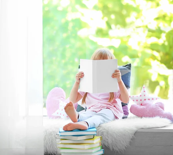 Vacker liten flicka med boken på fönsterbrädan — Stockfoto