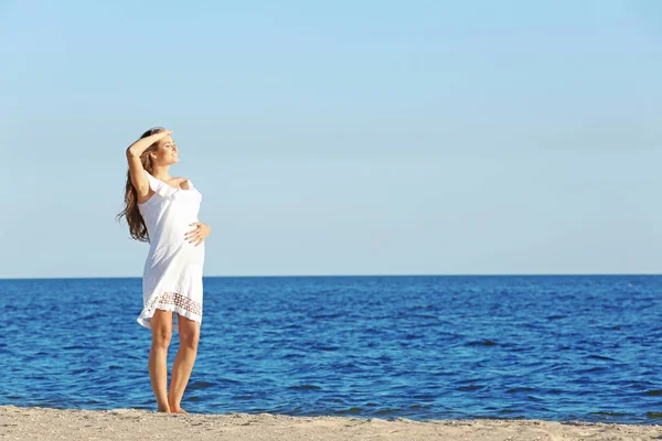 Mujer embarazada joven en vestido blanco de pie en la playa —  Fotos de Stock