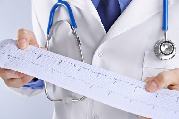 Male doctor holding cardiogram on light background, close up view