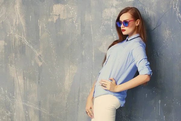 Jeune femme élégante en chemise bleue et lunettes de soleil sur fond gris — Photo