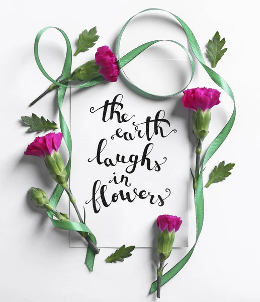 Inscriptie "De aarde lacht In bloemen" geschreven op papier met bloemen en lint op witte achtergrond — Stockfoto