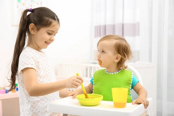 Nettes Mädchen füttert kleine Schwester im Hochstuhl im Babyzimmer — Stockfoto