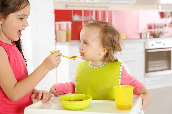 Linda chica alimentando a hermanita en silla alta en la cocina — Foto de Stock