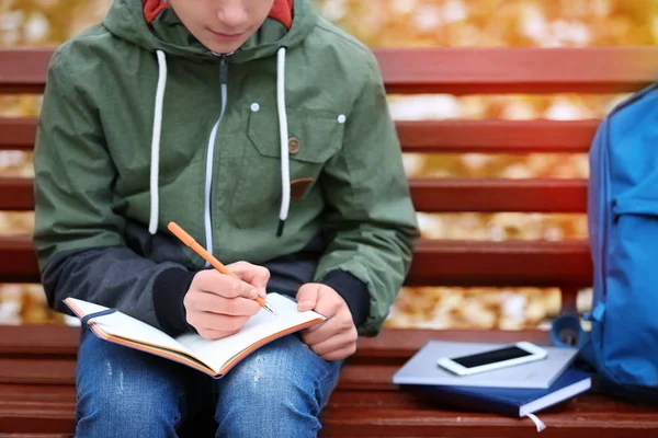 Teenager psaní v poznámkovém bloku při posezení na lavičce v podzimním parku, zblízka — Stock fotografie