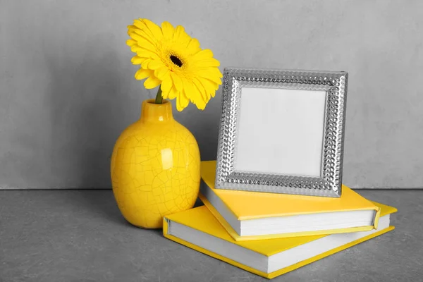 Yellow books, photo frame and vase with gerbera flower on grey background