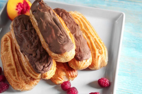 Deliciosos eclairs com chocolate, pêssego e framboesa na placa e mesa de madeira — Fotografia de Stock