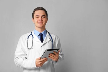 Young male doctor with tablet on grey wall background clipart