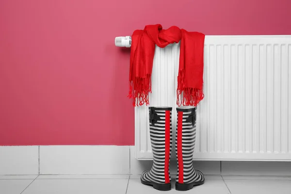 Gumboots and scarf near heating radiator on pink background — Stock Photo, Image
