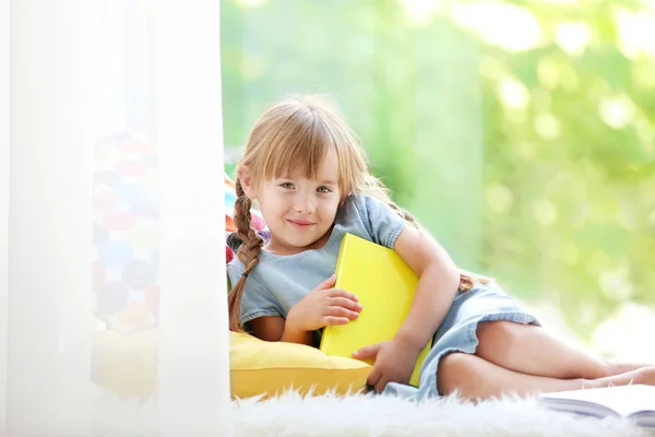 Lächelndes kleines Mädchen mit Buch auf Fensterbank liegend — Stockfoto