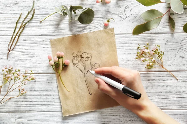 Kvinnlig hand ritning blomma på papper på vit trä bakgrund — Stockfoto