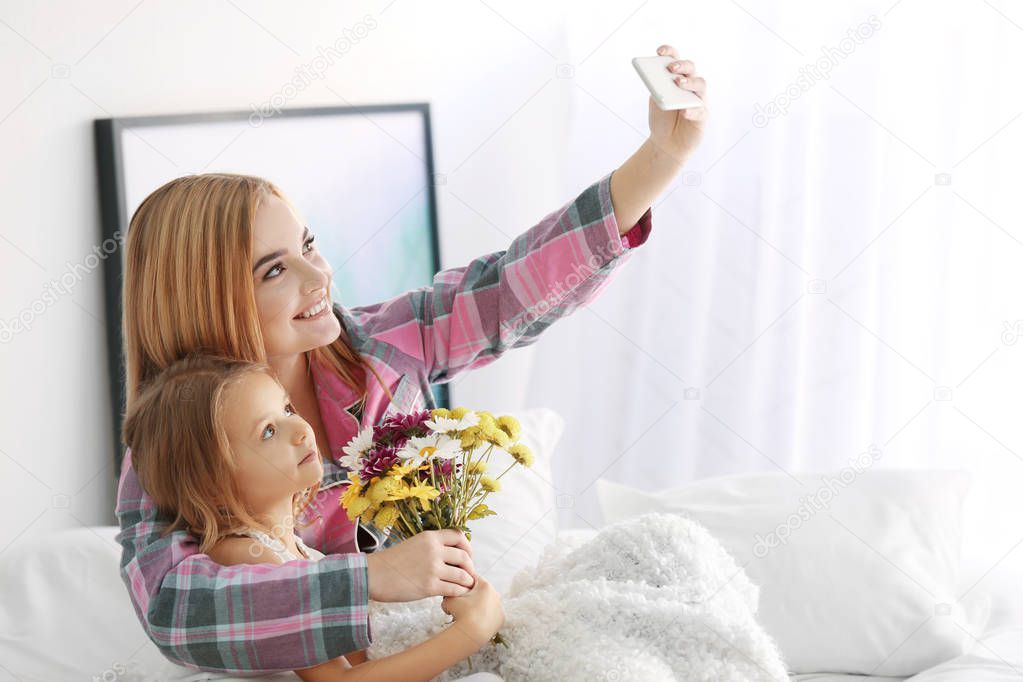 Beautiful young woman with daughter taking selfie while sitting on bed. Mother's day concept
