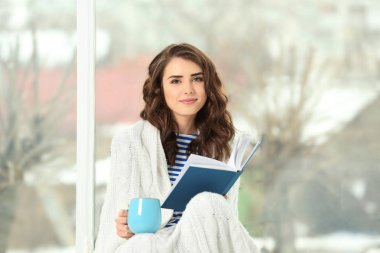 Güzel genç kadın kahve içme ve evde pencere yanında otururken kitap okuma