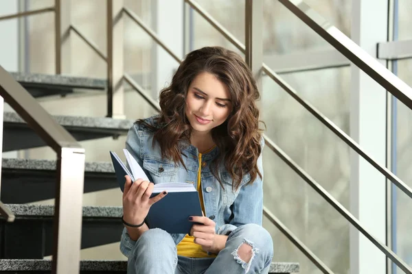 Mooie jonge vrouw lezen boek zittend op de trap in de buurt van venster — Stockfoto