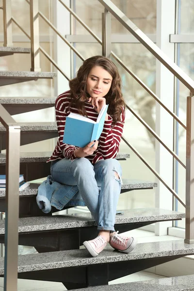 Όμορφη νεαρή γυναίκα που διαβάζει το βιβλίο ενώ κάθονται στα σκαλοπάτια κοντά σε παράθυρο — Φωτογραφία Αρχείου