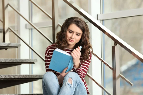 Beautiful young woman reading book while sitting on stairs near window — Stock Photo, Image