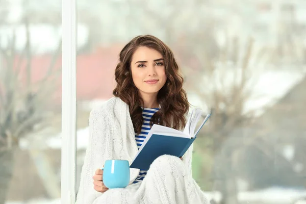 Belle jeune femme buvant du café et lisant un livre assis près de la fenêtre à la maison — Photo