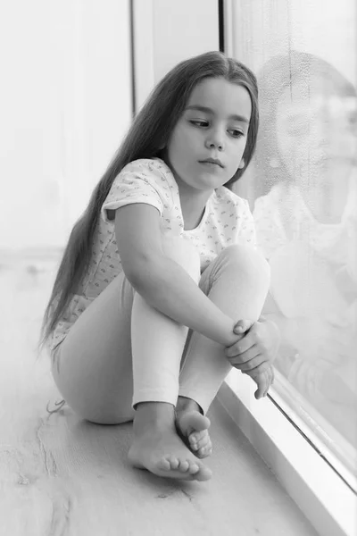 Маленькая грустная девочка сидит на подоконнике — стоковое фото