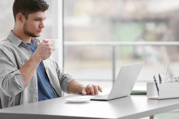 Beau jeune homme travaillant sur un ordinateur portable et buvant du café à la maison — Photo