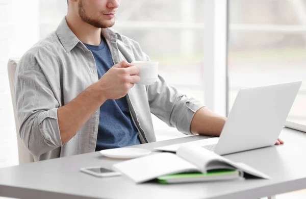 Jovem bonito trabalhando no laptop e bebendo café em casa — Fotografia de Stock