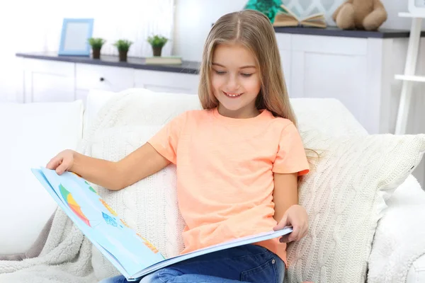 Nettes kleines Mädchen mit Buch sitzt zu Hause auf dem Sofa — Stockfoto